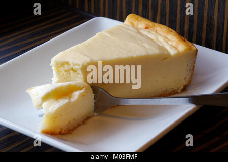 Plain cheesecake al forno con torta sulla forcella sul piatto di portata in ceramica bianca. Sfondo marrone. Foto Stock