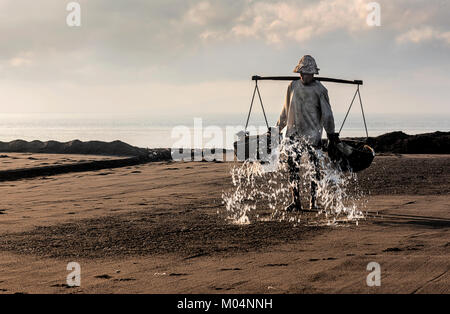 Sale indonesiano agricoltore di Kusamba sale villaggio minerario di Bali la raccolta di acqua di mare al mattino per la produzione di sale utilizzando il metodo tradizionale Foto Stock