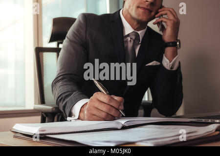 Imprenditore parlando al telefono cellulare mentre si lavora alla sua scrivania. Uomo seduto alla sua scrivania rendendo note e parlando al telefono cellulare. Foto Stock