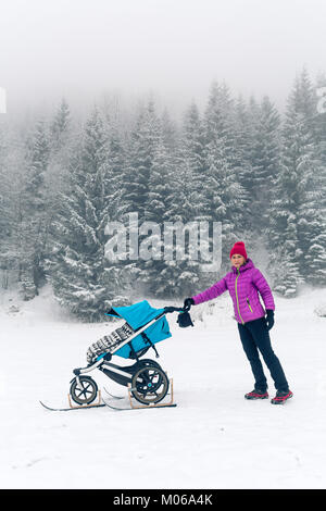 Madre con bambino passeggino godendo della maternità in inverno Foresta, Montagne Paesaggio. Il jogging o power walking donna con lo slittino pram in boschi. Beautif Foto Stock