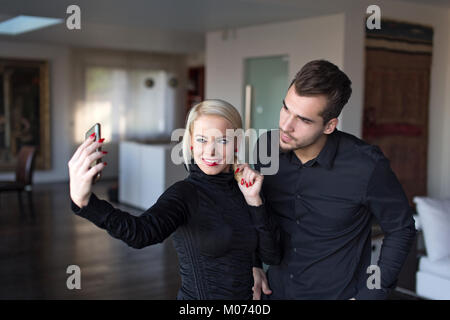 Felice giovane coppia elegante in abito nero tenendo selfie indoor Foto Stock