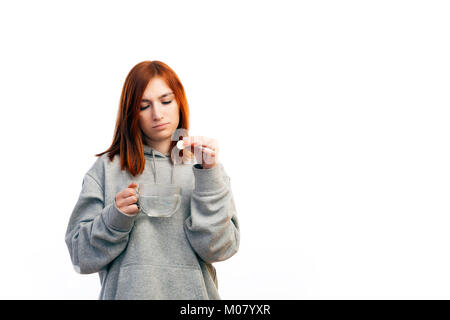 Un rosso giovane dai capelli donna in grigio di una felpa si ammala con un freddo e getta una grande pillola di vitamina A nell'acqua per ripristinare rapidamente su un bianco Foto Stock