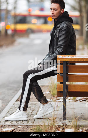 Abito moderno. Moda uomo sedersi sulla panchina e guardarsi intorno. In autunno e in inverno lo stile di abbigliamento. Giacca con sneakers bianco. Foto Stock