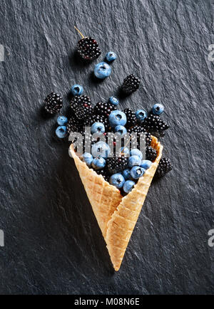 Esplosione di bacche diverse. Foto di mirtillo e blackberry nel cono di cialda su nero ardesia. Vista dall'alto. Alta risoluzione prodotto. Foto Stock