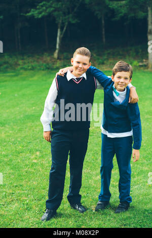 Due ragazzi, fratelli, scolari vestito in convenzionale con le braccia intorno a ogni altro di fronte a una foresta in un prato in estate. Foto Stock