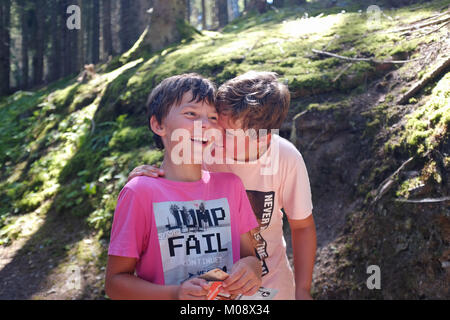 Due ragazzi, fratelli, passeggiate e ridere come uno di loro dice a un segreto con la sua mano su di una spalla di altri in una foresta. Foto Stock