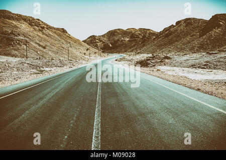 Un vuoto che la strada asfaltata che attraversa zone aride montagne nel Sultanato di Oman in Al Wusta Governate (vintage effetto della telecamera e il filtro) Foto Stock