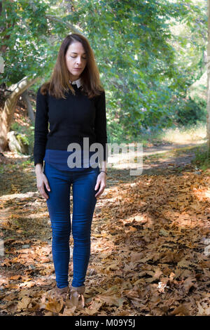 Un sottile donna matura, 46 anni, stando in piedi, corpo pieno, guardando verso il basso e le mani sulle gambe, natura dello sfondo. Foto Stock