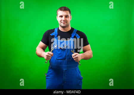 Un giovane uomo di costruzione builder in un T-shirt nera e blu globale di costruzione tiene nelle sue mani un nuovo strumento chiave di combinazione su una verde isolare Foto Stock