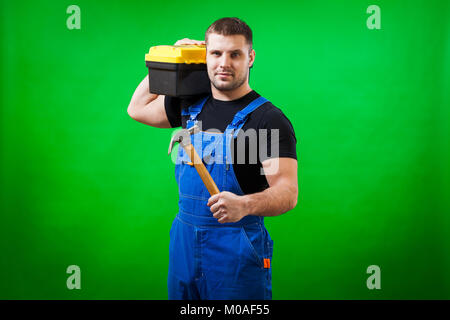 Un uomo forte builder in un T-shirt nera e blu tuta costruzione tiene un martello di legno e una scatola con strumenti di costruzione sulla sua spalla su una gr Foto Stock