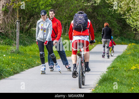 Un paio di pattinatori in linea e un ciclista condividono una pista ciclabile del parco. Persone attive che pattinano in linea su una pista ciclabile condivisa. Uomo che va in bicicletta all'aperto Foto Stock