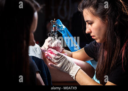 Una bella tatuaggio donna fa un tatuaggio di un piccolo uccello con vernice rossa con un tatuaggio macchina da una giovane donna sul suo hip Foto Stock