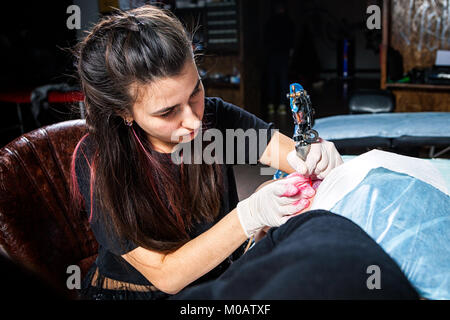 Una bella tatuaggio donna fa un tatuaggio di un piccolo uccello con vernice rossa con un tatuaggio macchina da una giovane donna sulla sua coscia in un salotto tattoo Foto Stock