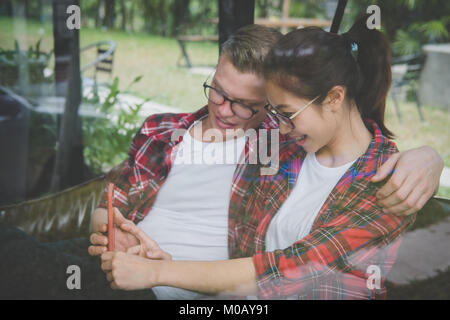 Coppia giovane in amore sorriso & look a smart phone. uomo caucasico & donna asiatica seduto sul divano di casa nei pressi di finestra. ragazzo e ragazza risalente al cafe. Foto Stock