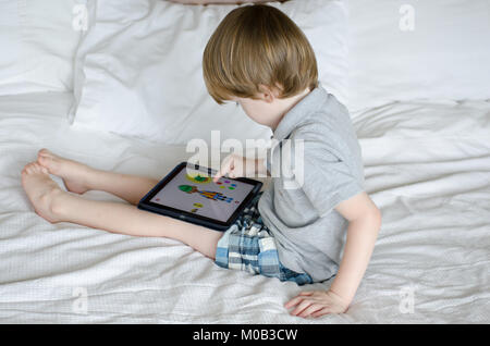 Little Boy seduta sul letto e giocare su un iPad Foto Stock