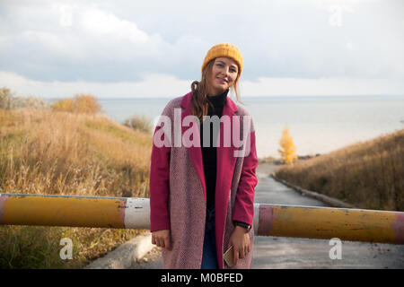 Un buio dai capelli donna in un giallo berretto lavorato a maglia, un cappotto rosa è in piedi sulla strada vicino al campo di grano in una giornata autunnale, sullo sfondo di una betulla Foto Stock