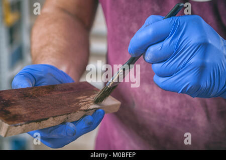 Close-up di un giovane maschio falegname di costruzione in viola calcio lavoro protettivi e guanti monouso dipinge un bar in legno con un pennello in un oscuro Foto Stock