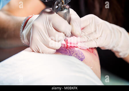 Close-up di una giovane donna tattoo master in guanti sterili rende un tatuaggio sulla coscia con vernici di colore rosso Foto Stock