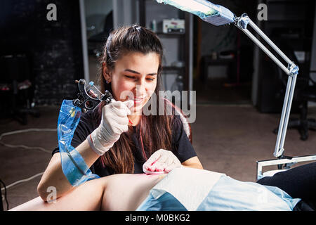 Giovane donna tattoo artist sorridente e facendo con Tatuaggio Tattoo Machine sotto la luce della lampada in officina scuro Foto Stock