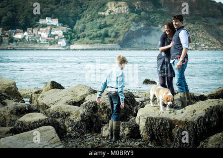 Ragazzo giocando sulle rocce con i suoi genitori e il cane di famiglia. Famiglia godendo le vacanze sulla costa del Regno Unito, mare nella baia di Runswick, Yorkshire. Foto Stock