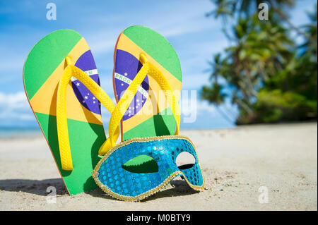 Bandiera Brasile flip-flop e luminosamente sequined maschera di Carnevale in appoggio insieme nella sabbia sulla riva di un orlata di palme spiaggia brasiliana Foto Stock