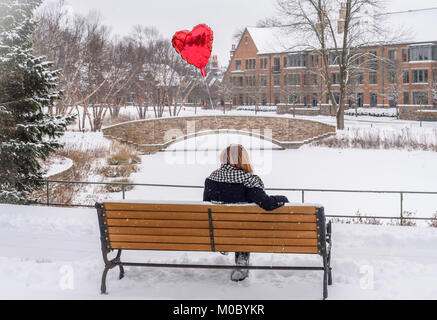 Donna seduta al di fuori su una panchina nel parco azienda rosso a forma di cuore ballood in luce la caduta di neve Foto Stock