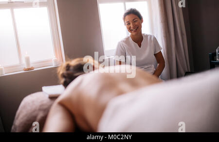 Donna asiatica massaggiatrice sorridente e parlando con la femmina sdraiato sul lettino da massaggio nella spa salone. Massaggio femmina terapista di parlare al cliente al centro benessere Foto Stock