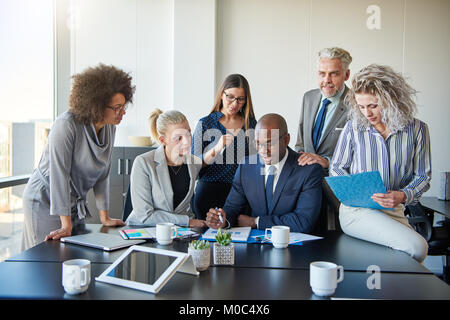 Gruppo di imprenditori focalizzati in piedi intorno al loro manager seduto in un ufficio tavolo riunioni rivedendo i grafici e documenti Foto Stock