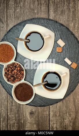 Bevande, acqua potabile, energia e caffeina concept-due tazze di caffè, caffè in grani, macinato di caffè e caffè istantaneo al buio su un supporto di ardesia. Foto Stock