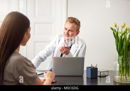 Giovani sorridente cosmetologo specialista uomo seduto a un computer portatile in ufficio e comunicante con una femmina di client. Foto Stock