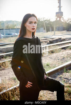 Moda shot: ritratto della bella ragazza rock (modello informale) in tunica e pantaloni in piedi alla ferrovia (zona industriale) Foto Stock