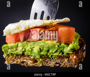 Morso dimensioni pezzo di avocado toast con pomodoro e un uovo fritto su una forcella. Isolato su sfondo nero Foto Stock