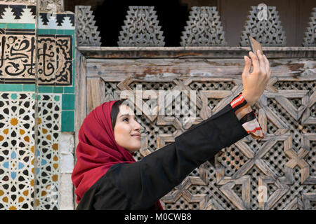 La donna araba in abbigliamento tradizionale tenendo selfie con telefono cellulare nella parte anteriore della parete con testo dal Corano Foto Stock