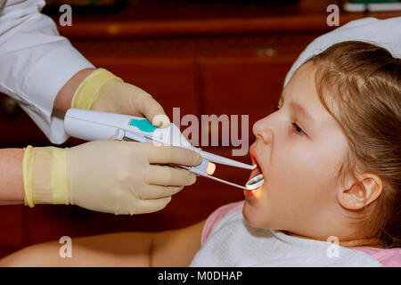 Baby girl dentista esamina i denti del paziente allegro bambino con ricci capelli rossi. ragazza sorridente in poltrona del dentista. bambino bocca aperta in Foto Stock