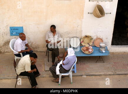 Quattro uomini seduti e chat sul ciglio della strada accanto a una tabella con un paio di articoli per la vendita, Douz, Kebili distretto, Tunisia Foto Stock