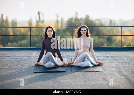 Due belle donne eseguire la posa meditativa gomukhasana sul tetto all'aperto. Giovani ragazze attraenti sedersi tranquillamente in una posa di rilassamento. Concentrazione, h Foto Stock