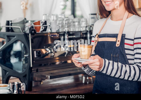 Asia donna barista indossare jean grembiule tenendo calda tazza di caffè servito al cliente con volto sorridente al banco bar,ristorante Cafe concetto di servizio.cameriera Foto Stock