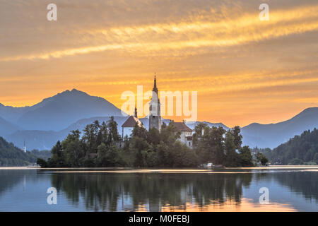 Il famoso Lago di Bled con la chiesa di Santa Maria e le montagne sullo sfondo arancione sotto il cielo mattutino, Slovenia, Europa Foto Stock