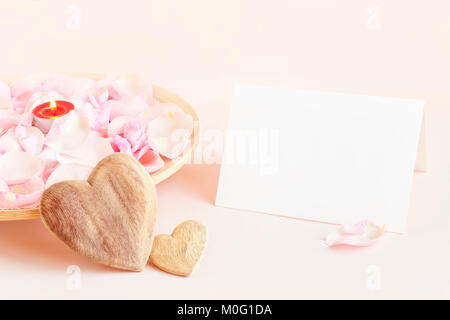 Vuoto carta bianca su sfondo rosa con piastra in legno riempita con il colore rosa dei petali di rosa e la candela e due cuori di legno per San Valentino Foto Stock