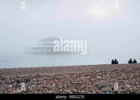 La gente seduta sulla spiaggia guardando il Molo Ovest di Brighton, la più popolare destinazione balneare nel Regno Unito per i turisti stranieri. Foto Stock