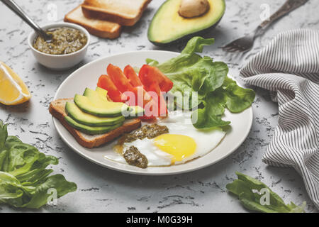 Dieta sana prima colazione. toast con fette di avocado, Uovo fritto, pomodori, crescione, pesto su una piastra su un sfondo di calcestruzzo Foto Stock