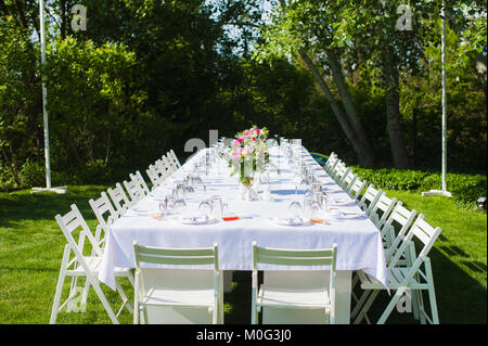 Stile rustico tavolo con tovaglia bianca e decorazioni floreali nel bellissimo giardino. Bianco e lungo matrimonio arte tavolo con sedie in legno, posate, Foto Stock