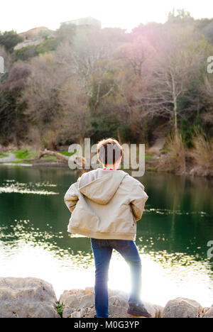 Ripresa posteriore di una decina di anni di vecchio ragazzo con giacca a vento e jeans in piedi sulle rocce guardando fuori attraverso un tranquillo fiume che scorre nella valle dell'Herault, Francia Foto Stock