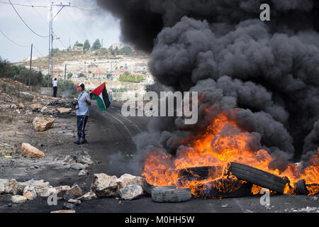 L'uomo con la bandiera palestinese in piedi vicino a bruciare pneumatici venerdì di rage. Kafr Qaddum, Palestina. © Antonio Ciufo Foto Stock