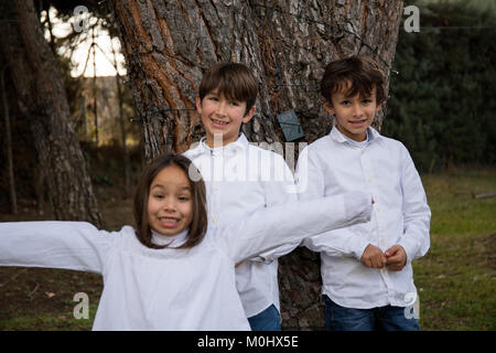 Due fratelli gemelli e una sorella più giovane in posa vicino ad un albero nel giardino della casa Foto Stock