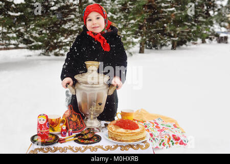 La ragazza del bambino in una pelliccia e in una sciarpa in stile russo tenendo un grande samovar nelle mani di frittelle con caviale rosso Foto Stock