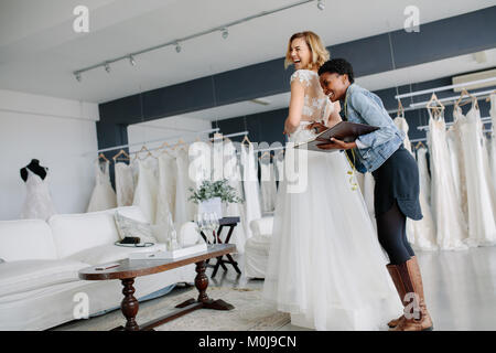 Cerca femmina in abito da sposa con donne assistant in nuziale negozio di usura. Donna sorridente che indossa il suo abito da sposa con abito da sposa designer in bridal f Foto Stock