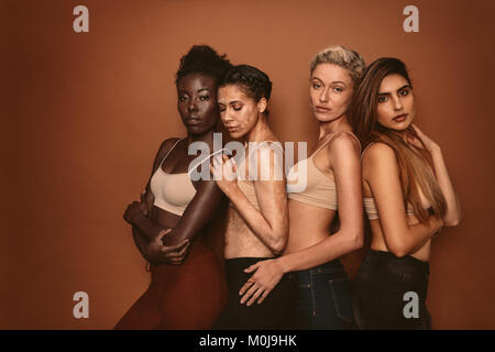 Il gruppo di quattro giovani diverse ragazze su sfondo marrone. Modelli femmina con skin differenti in piedi insieme e guardando la fotocamera. Foto Stock