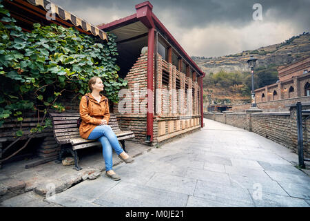 Tourist donna in giacca marrone seduto sul banco vicino a pubblico bagno solforico quartiere centrale di Tbilisi, Georgia Foto Stock