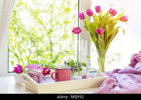 Accogliente casa concetto. Viola tulipani freschi in vaso di vetro. Amaretti in un barattolo di vetro. Tazza di tè caldo. Vassoio bianco Coperta di Lilla sul davanzale. Sunshine. C Foto Stock